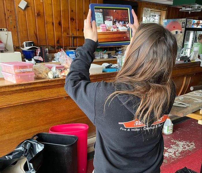 a female with brown hair wearing servpro uniform taking pictures of fire damaged content from a bar fire 