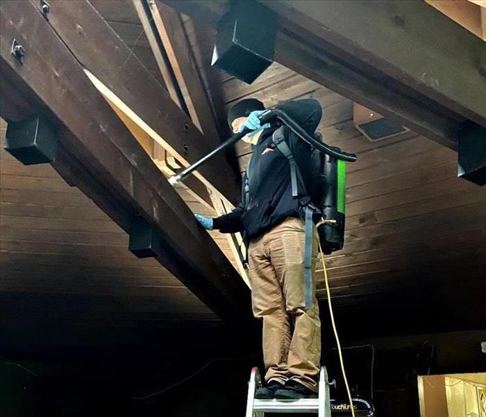 a man with khaki pants and black SERVPRO top standing on a ladder with a Hepa Vacuum cleaning soot particles from a fire job 